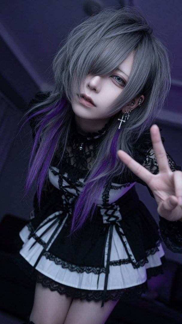 A person with silver and purple hair, wearing a black and white gothic-style dress, makes a peace sign.