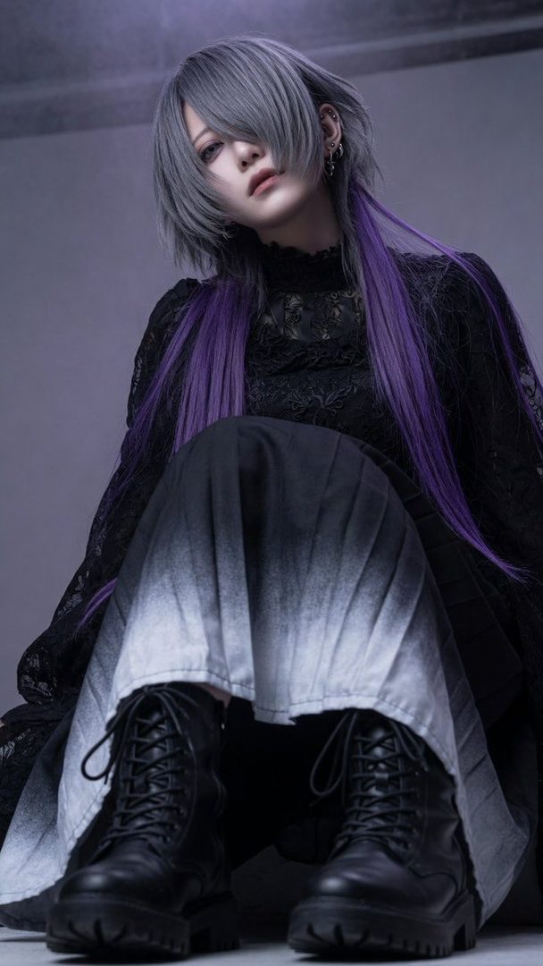 A person with silver and purple hair, wearing a black lace dress and combat boots, poses indoors with a cool expression.