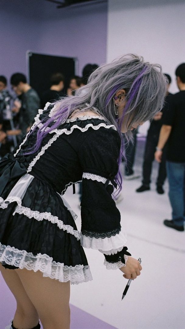 A person with long silver and purple hair, wearing a black and white frilly dress, is bending down indoors while holding a pen.