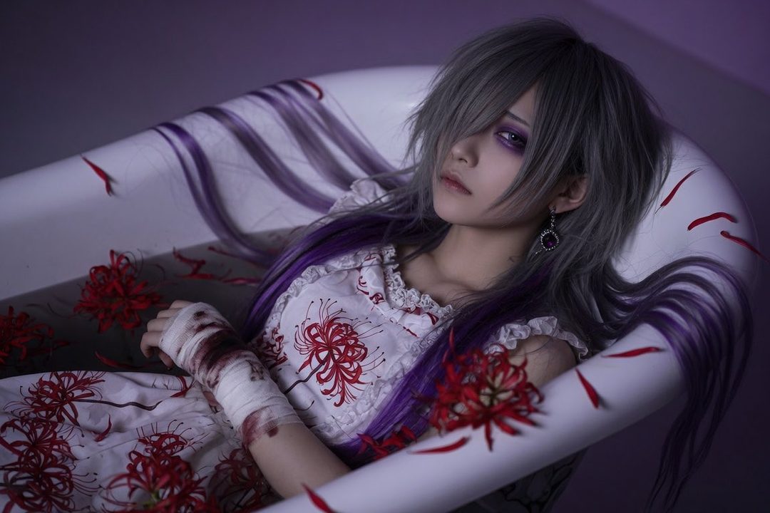 A person with silver and purple hair, wearing a white dress with red spider lily patterns, lies in a bathtub with red petals.