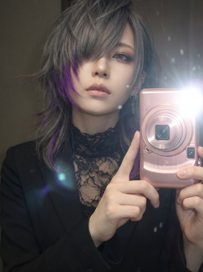 A person with silver and purple hair takes a selfie with a pink digital camera, wearing a black suit jacket over a lace top.