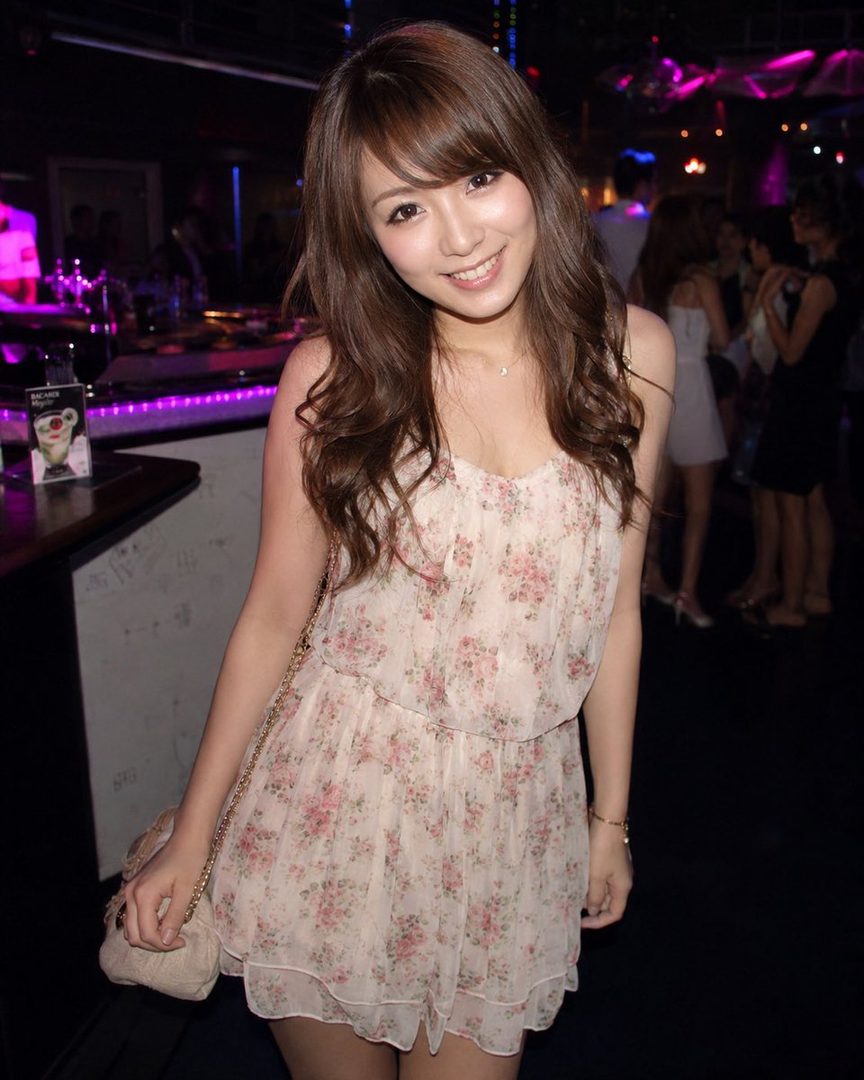A young woman with long brown wavy hair smiles brightly while wearing a floral dress in a dimly lit nightclub.
