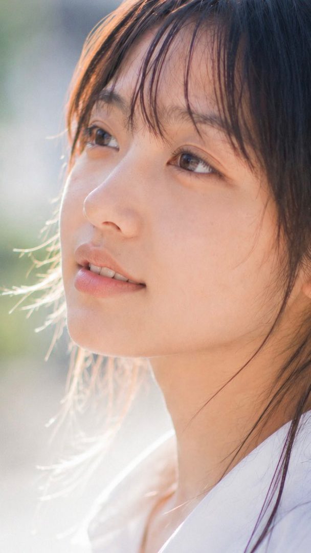 A close-up portrait of a young woman with dark hair and bangs, looking upwards with a serene expression in soft daylight.