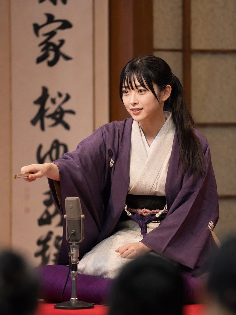 A young woman with black hair tied in a ponytail and bangs, wearing a purple kimono, sits indoors with a microphone in front of her.