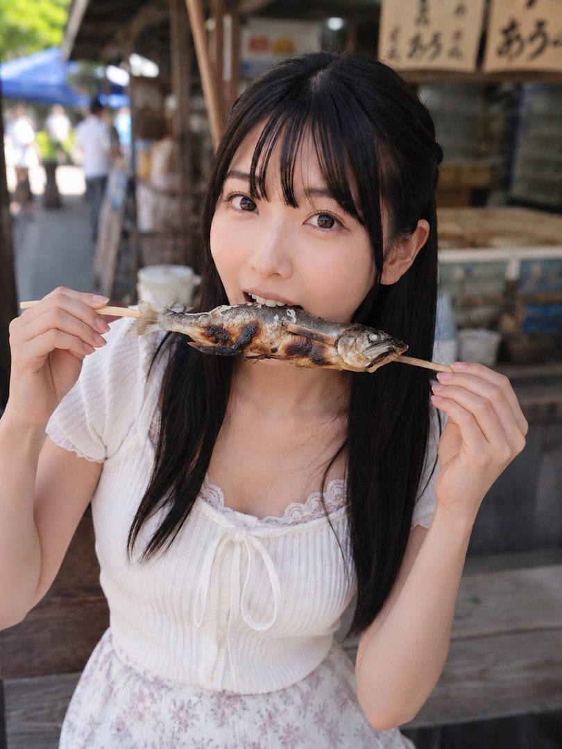 A young woman with long black hair and bangs holds a grilled fish on a stick to her mouth, looking at the camera outdoors.