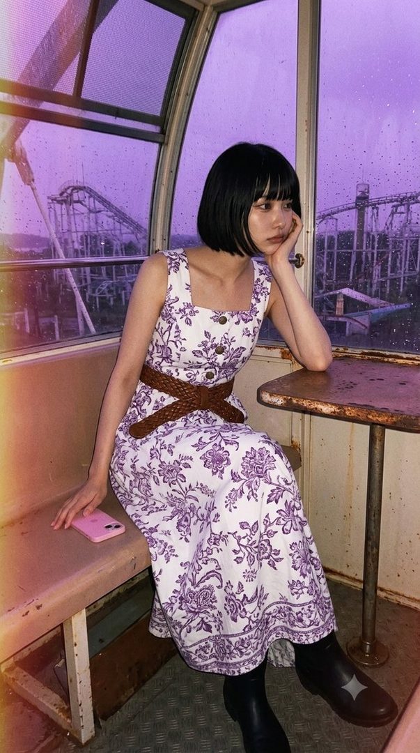 A woman with short black hair and bangs wears a white and purple floral dress, sitting inside a Ferris wheel cabin with a purple-hued amusement park visible outside.
