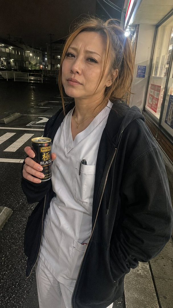 A woman in a nurse's uniform and black hoodie stands outdoors at night, holding a can of coffee.