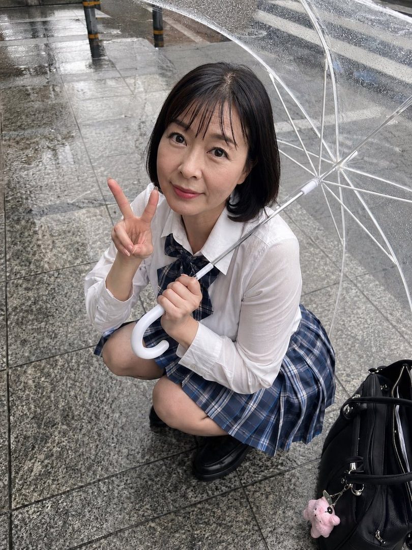 A woman in a school uniform crouches on a wet street, holding a clear umbrella and making a peace sign.