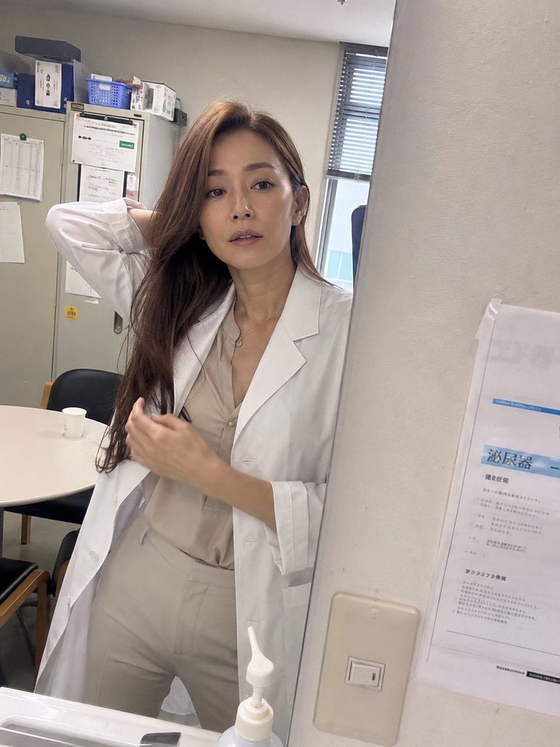 A woman with long brown hair wearing a white lab coat and beige top and pants, standing in an office or clinic setting.