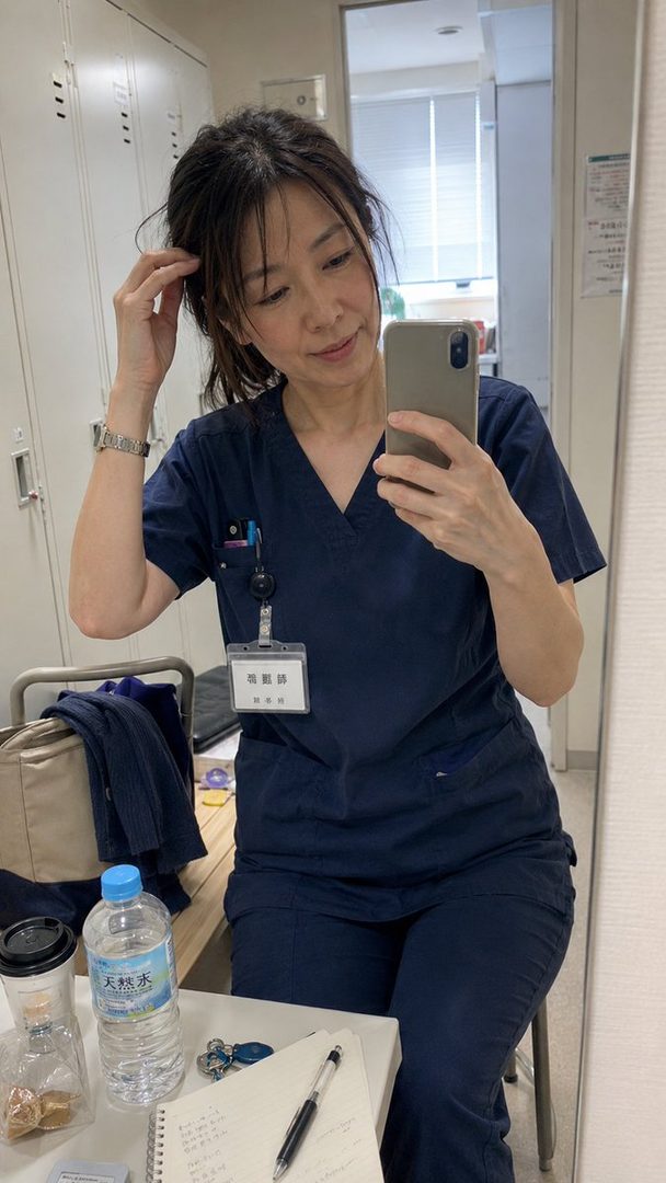 A woman in blue scrubs takes a mirror selfie in what appears to be a locker room or office.