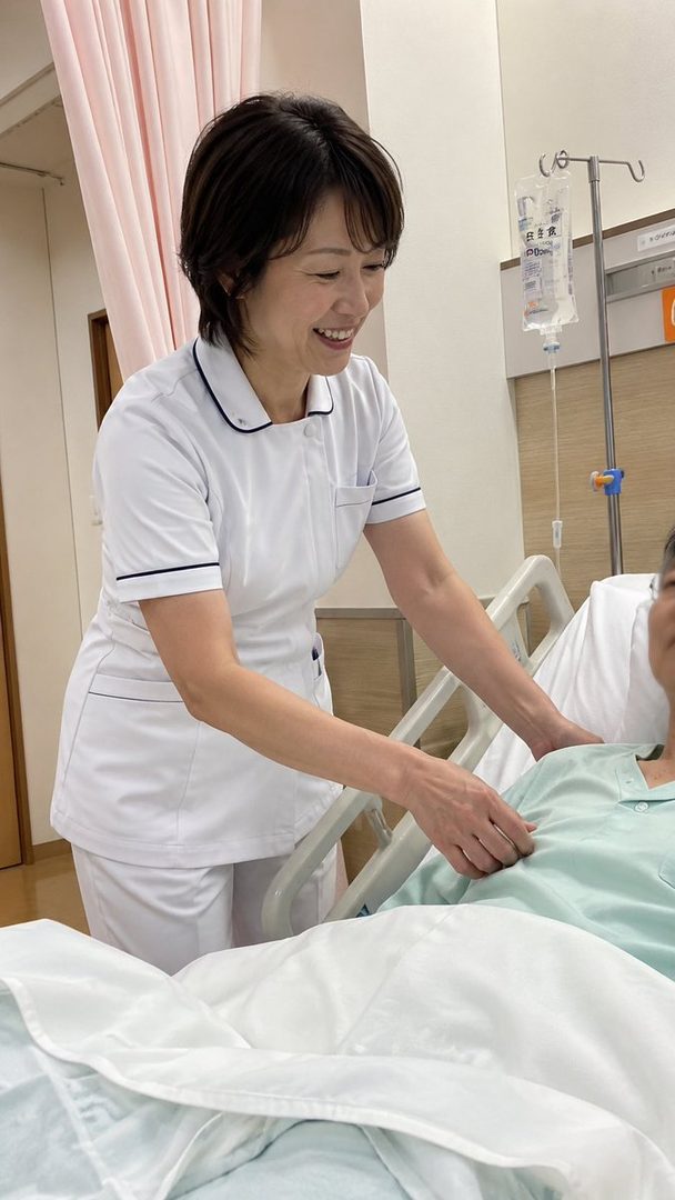 A smiling nurse in a white uniform attends to a patient in a hospital room.
