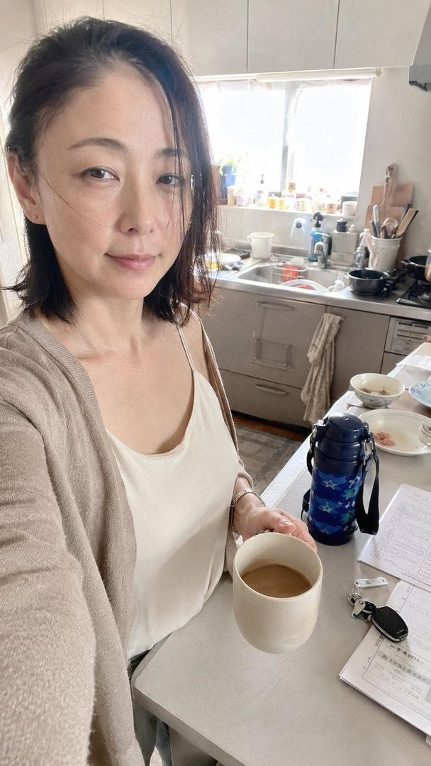 A woman with short dark hair takes a selfie in her kitchen, holding a cup of coffee.