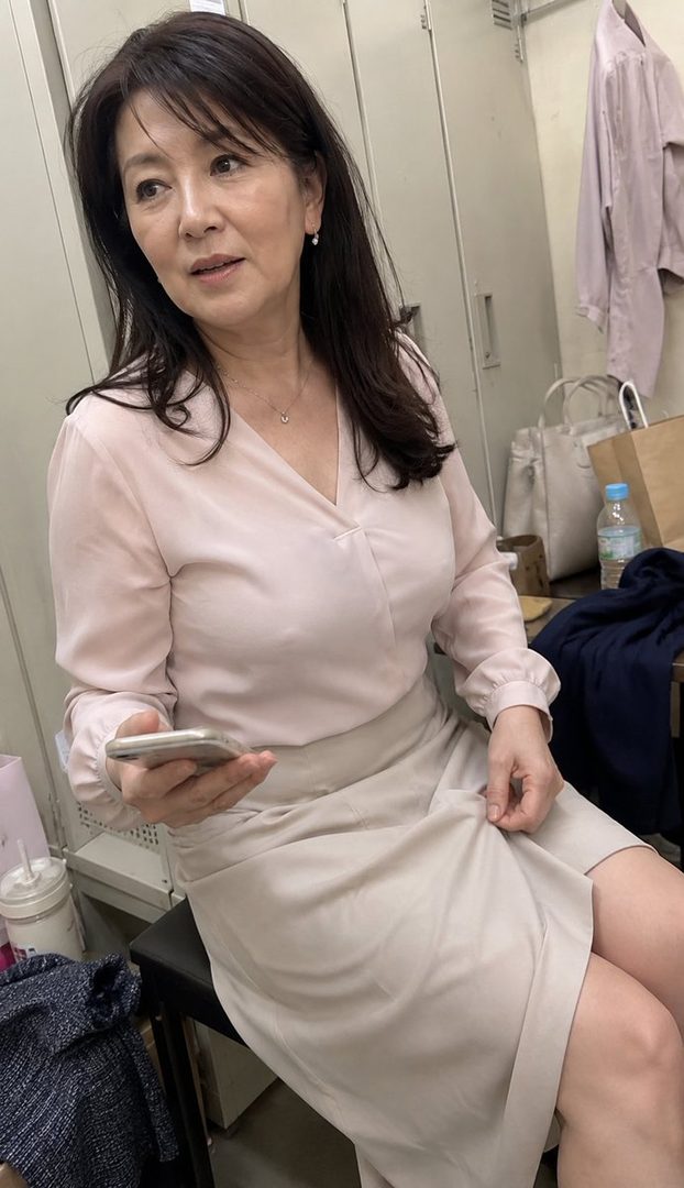 A mature woman with long brown hair and a light pink blouse sits indoors, holding a smartphone.