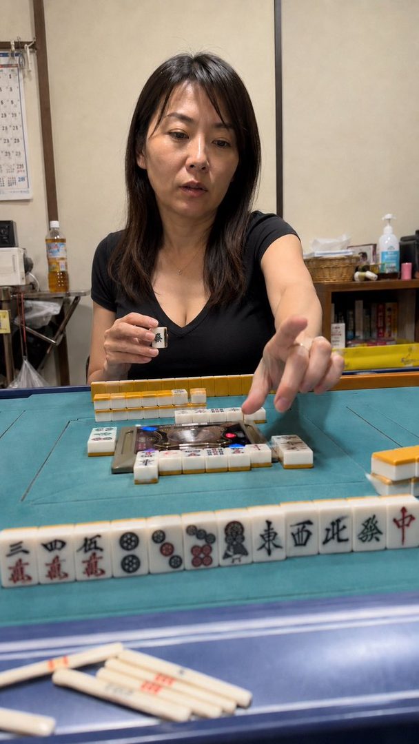 A photorealistic AI-generated image of a woman with long black hair playing mahjong indoors.