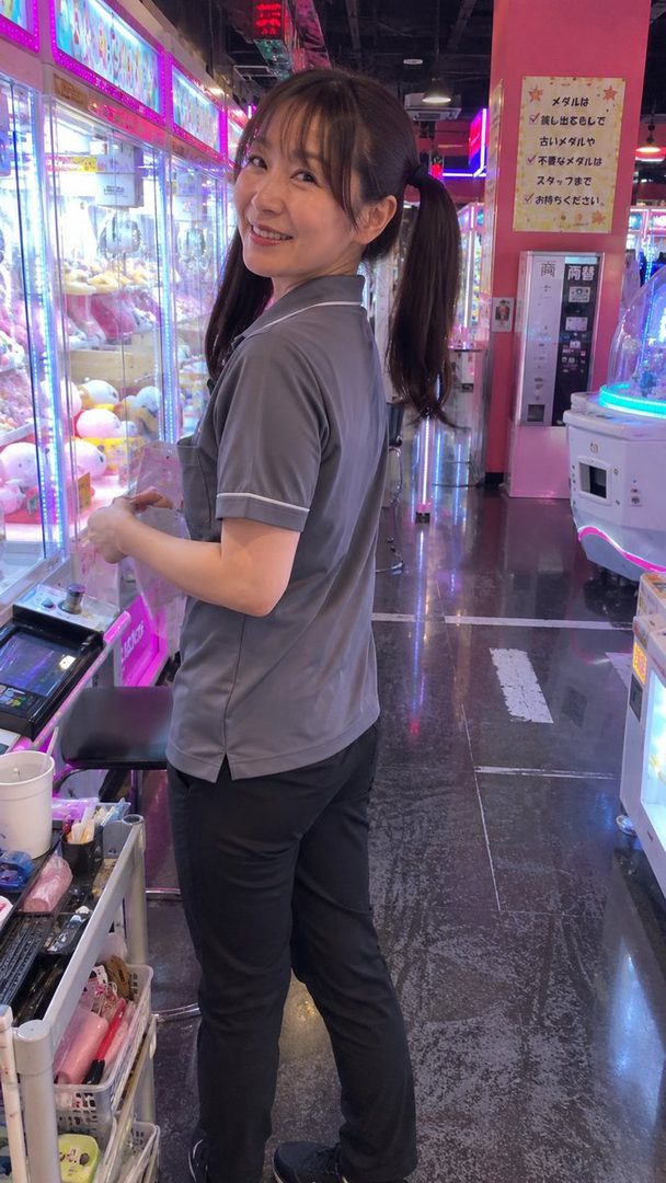A smiling woman with brown hair in pigtails, wearing a grey polo shirt and black pants, stands in an arcade.