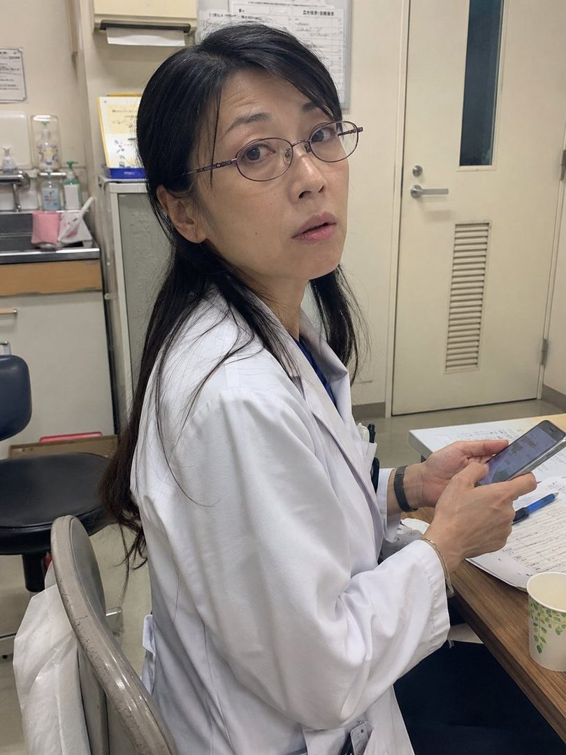 A woman in a white lab coat and glasses looks over her shoulder while holding a smartphone in an indoor setting.