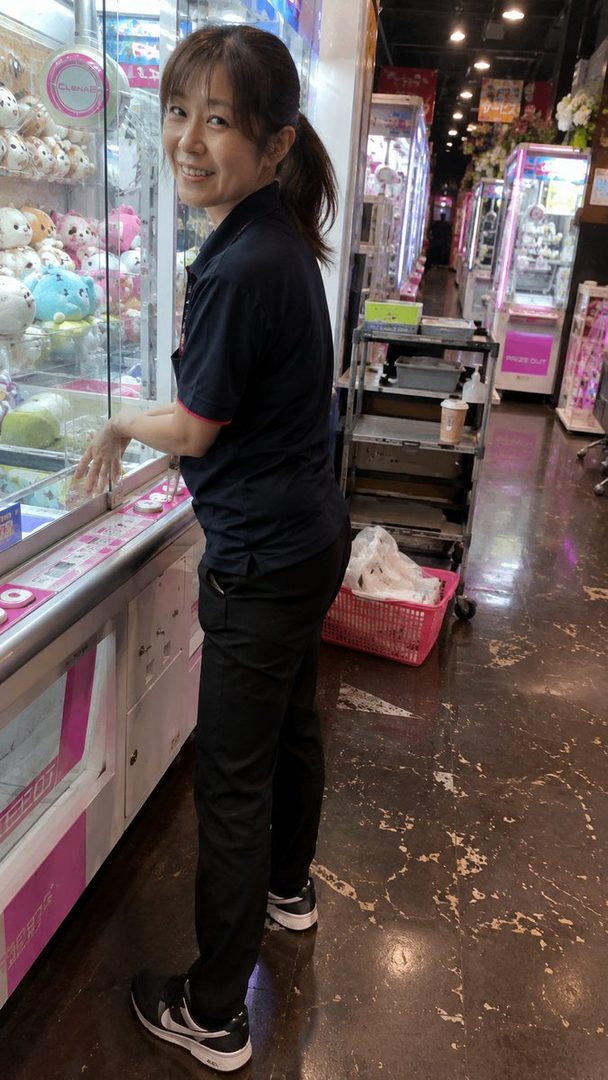 A smiling Japanese woman with a ponytail stands next to a claw machine in an arcade.