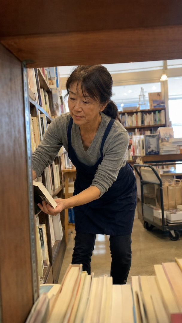 A woman with brown hair in a ponytail, wearing a grey long-sleeved shirt and a dark apron, is organizing books on a shelf in a bookstore.