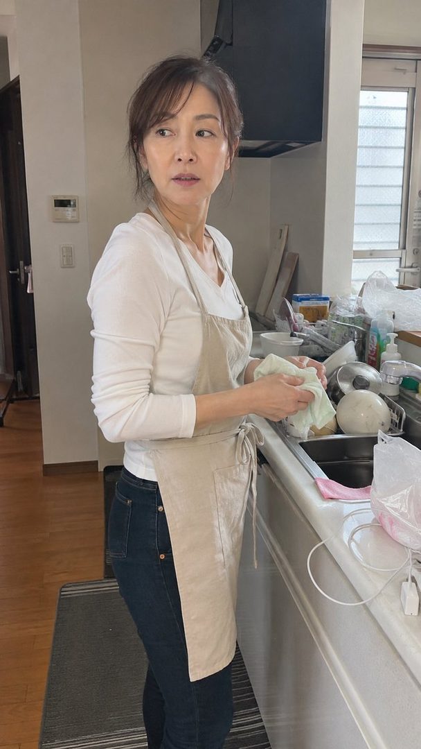 A Japanese woman in a white long-sleeve shirt and apron is washing dishes in a kitchen.