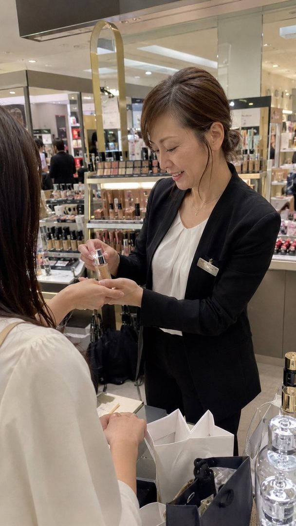 A Japanese beauty consultant in a black suit and white blouse is smiling while demonstrating a product to a customer in a department store.