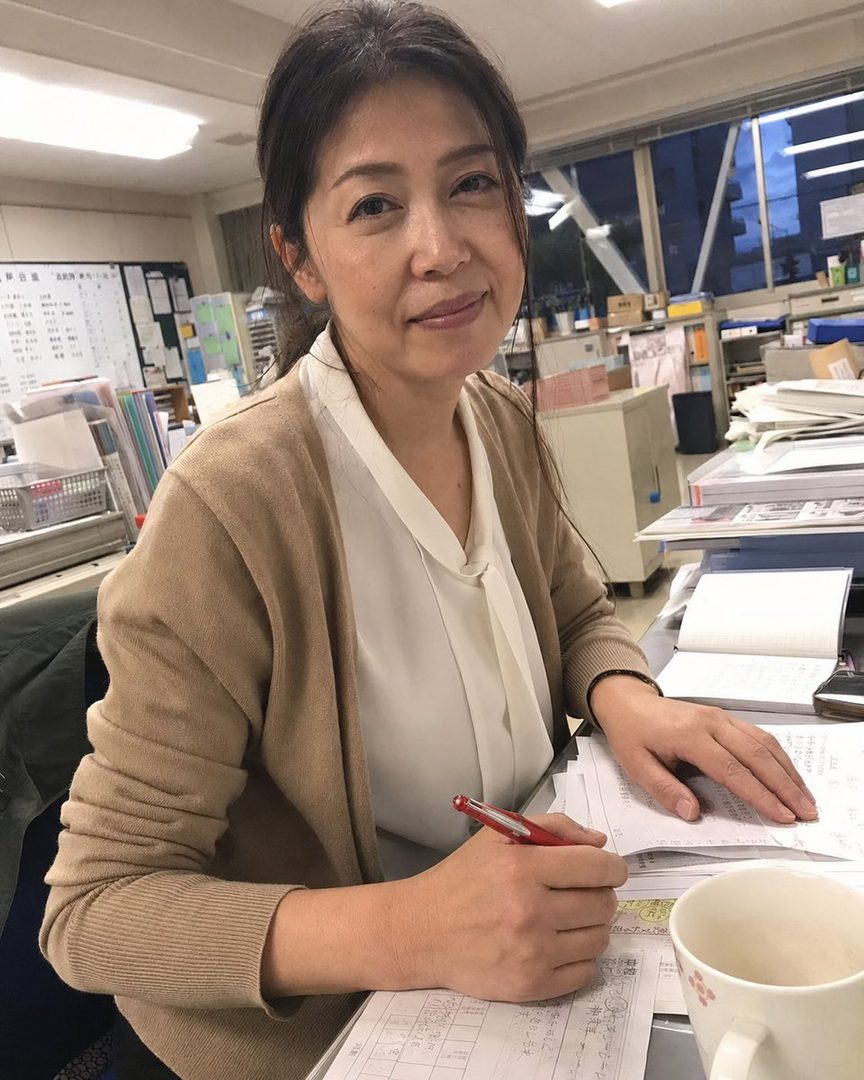 A Japanese woman in a brown cardigan and white blouse is working at an office desk, holding a red pen.