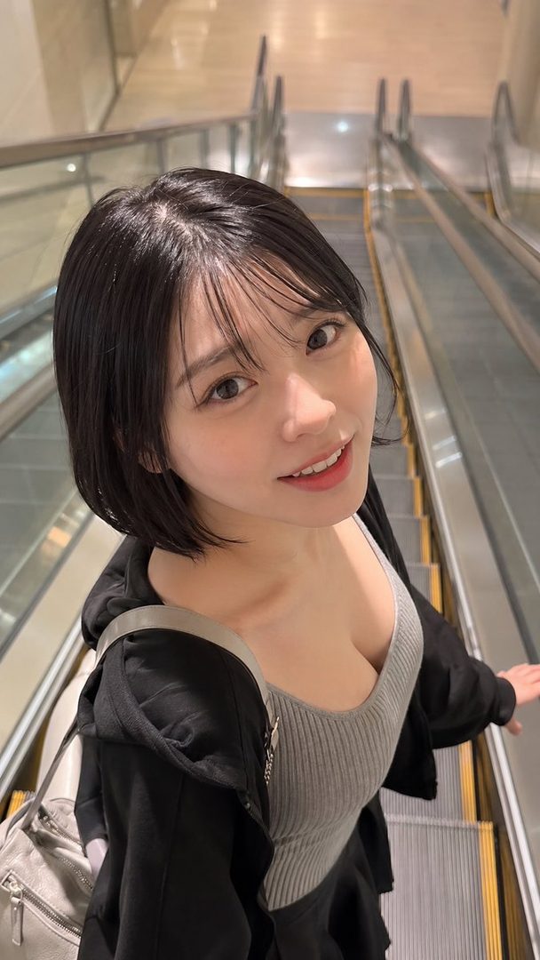 A young woman with short black hair and bangs smiles at the camera while riding an escalator indoors.