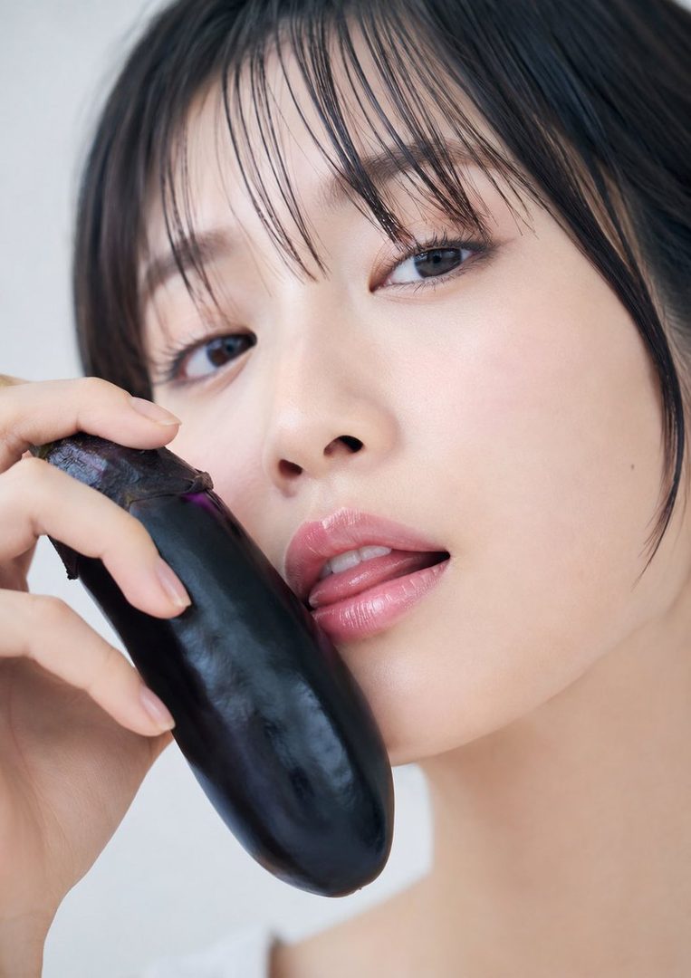 A close-up portrait of a woman with black hair and bangs, holding an eggplant near her mouth with her tongue slightly out.
