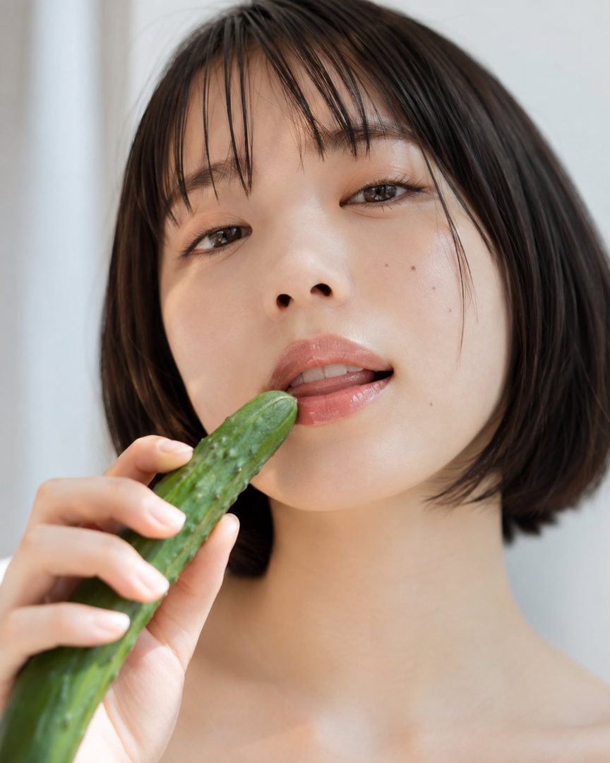 A woman with short black hair and bangs holds a cucumber to her lips in a suggestive manner.