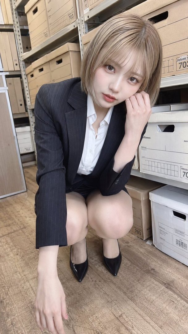 A young woman with blonde bob hair and a pinstripe suit crouches in an office setting with shelves of boxes.