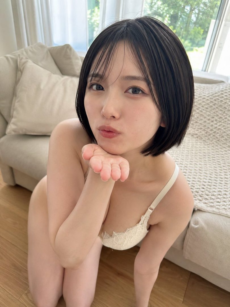 A young woman with short black hair and a white lace bra kneels on a wooden floor, blowing a kiss towards the viewer.