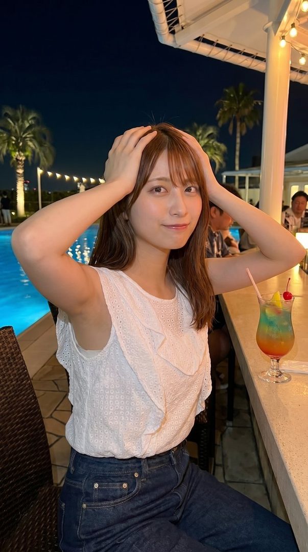 A young woman with brown hair and bangs poses outdoors at night, wearing a white top and jeans, with a colorful drink on the bar beside her.