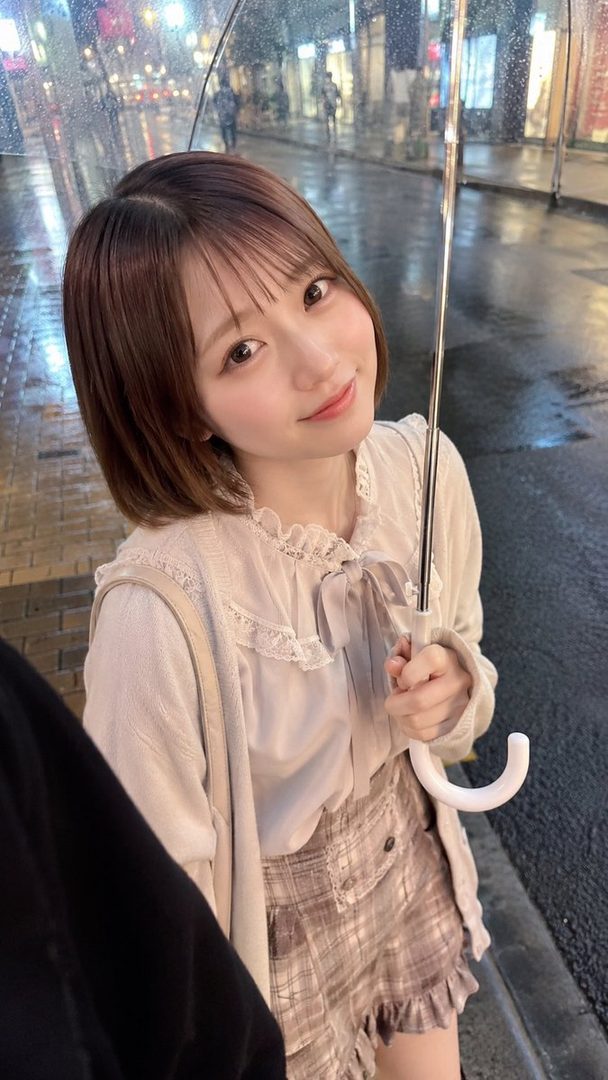 A cute young woman with short brown hair smiles while holding an umbrella on a rainy city street at night.
