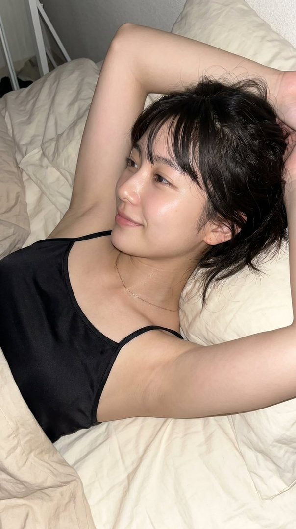 A young woman with short black hair and bangs lies in bed, wearing a black camisole, looking to the side with a gentle smile.