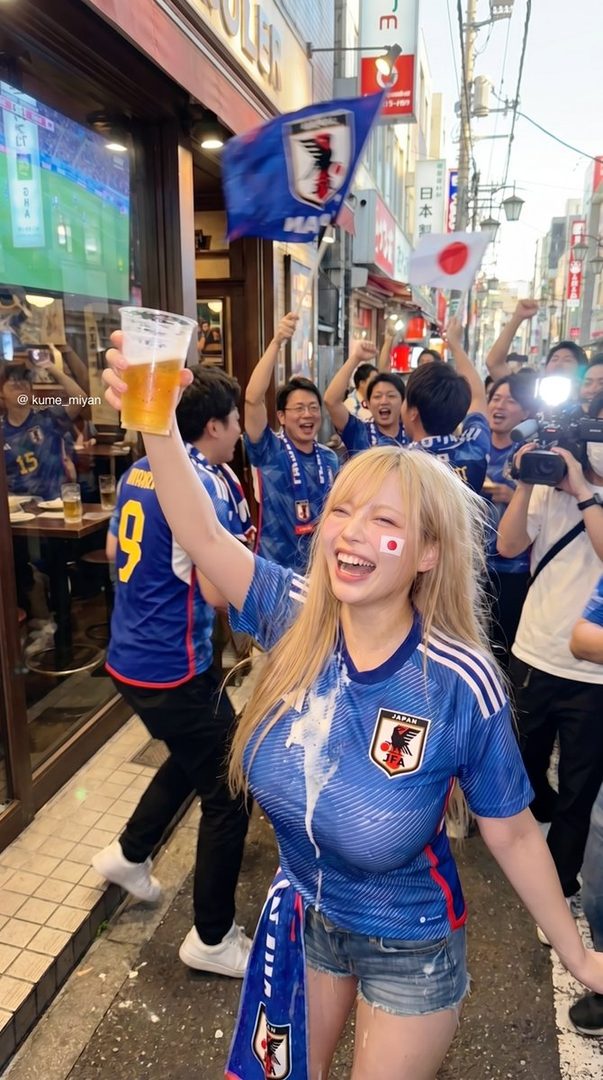 A cheerful woman with blonde hair in a Japan soccer jersey celebrates on the street, holding a beer.