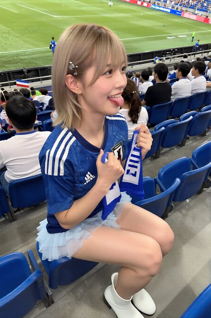 A young woman with blonde hair wearing a blue soccer jersey and a light blue ruffled skirt, sticking her tongue out playfully while sitting in a stadium.