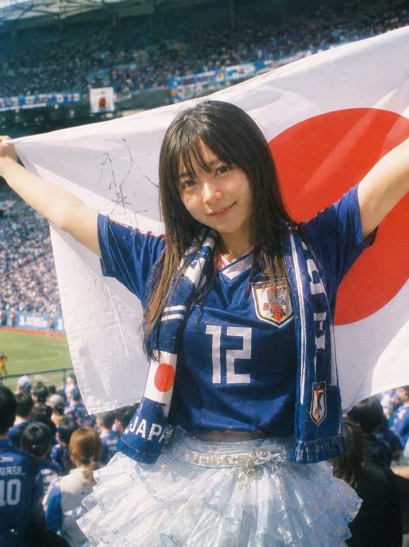 A young woman in a Japan national soccer team jersey and scarf holds a Japanese flag at a stadium.