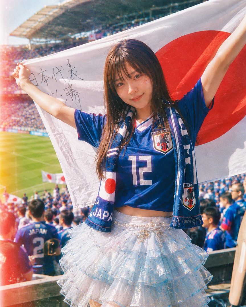 A young woman in a blue Japan soccer jersey and a frilly skirt holds a Japanese flag at a stadium.