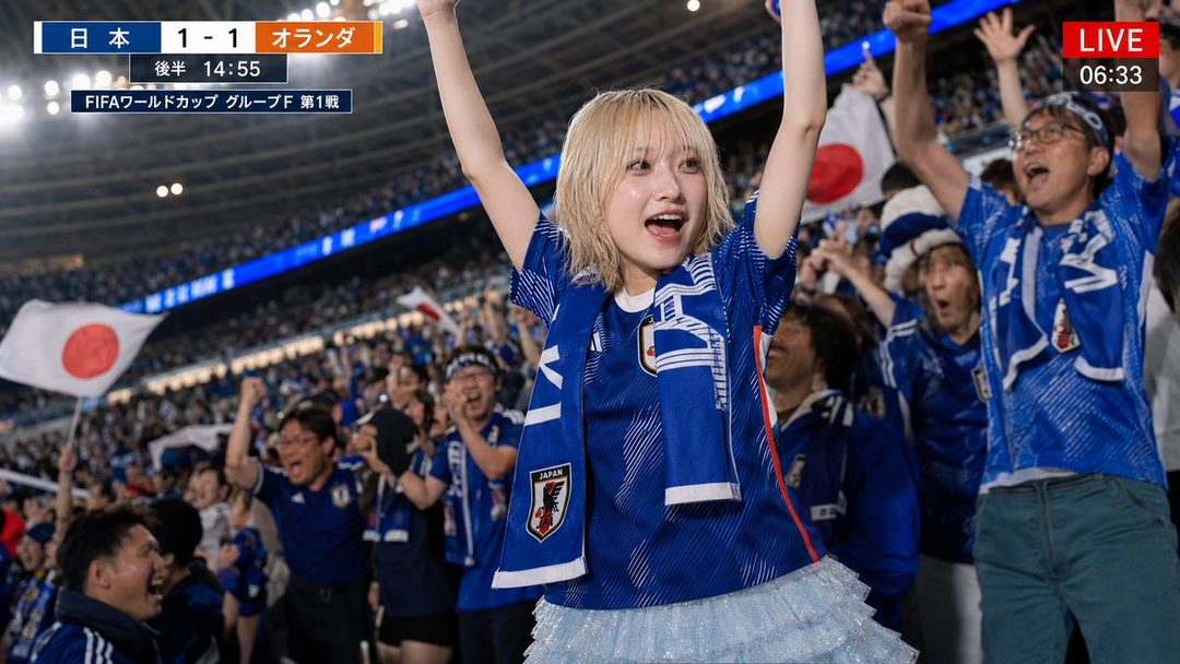 A young woman with blonde hair wearing a Japan national football team jersey and scarf, cheering enthusiastically at a World Cup match.