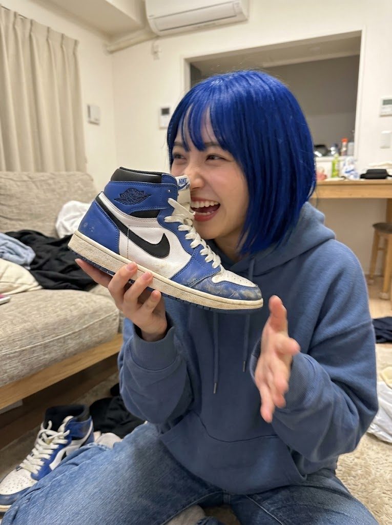 A young woman with blue hair and a blue hoodie is sitting on the floor, holding up a blue and white sneaker and smiling.