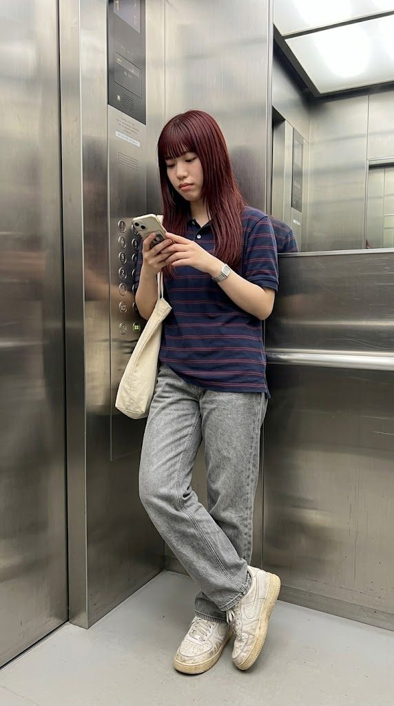 A young woman with red hair and bangs, wearing a striped polo shirt and jeans, stands in an elevator looking at her phone.