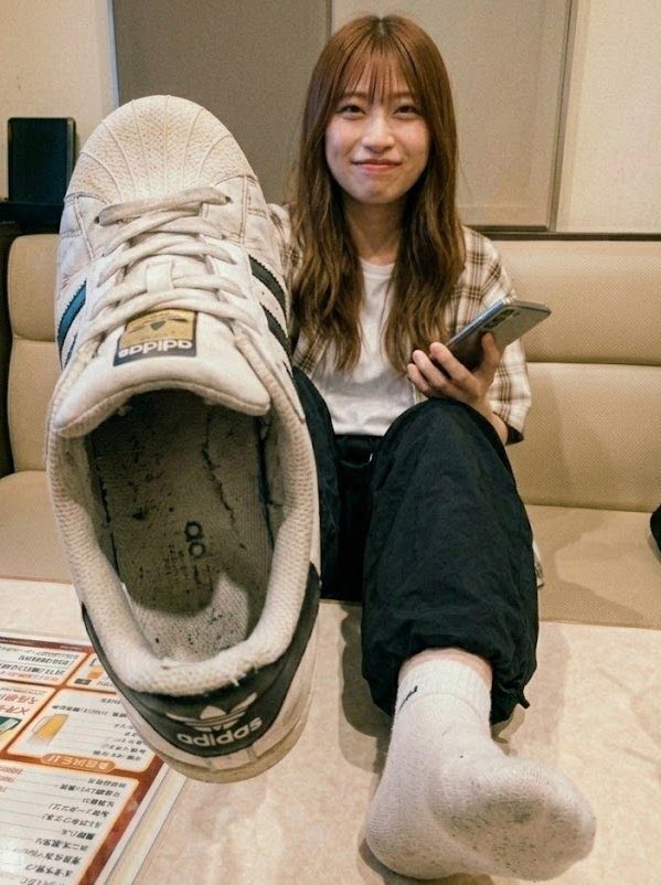 A young woman with long brown hair and bangs, wearing a plaid shirt and dark pants, holds up a large white Adidas shoe while sitting indoors.