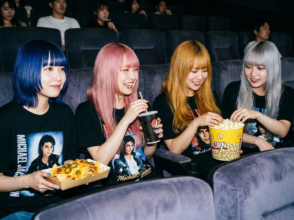 Four young women with colorful hair are enjoying snacks and drinks while watching a movie in a cinema.