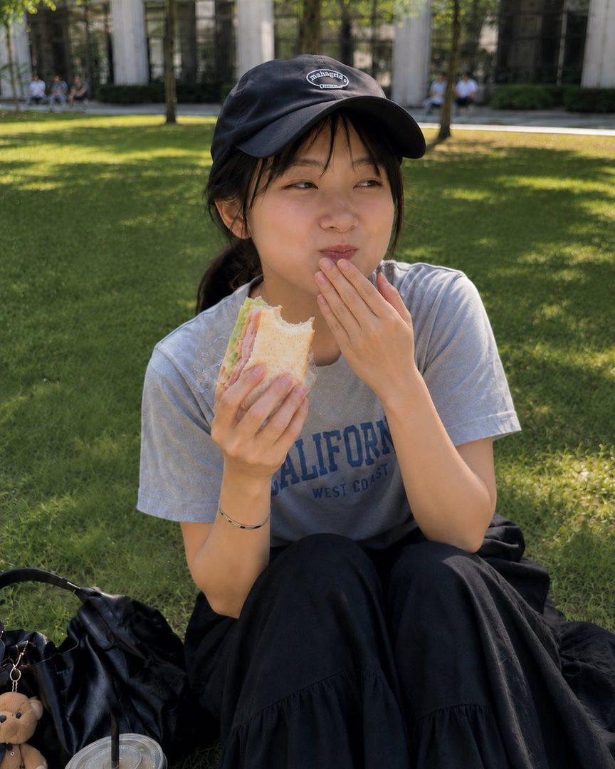 A young woman in a black cap and grey t-shirt is sitting on the grass, eating a sandwich.