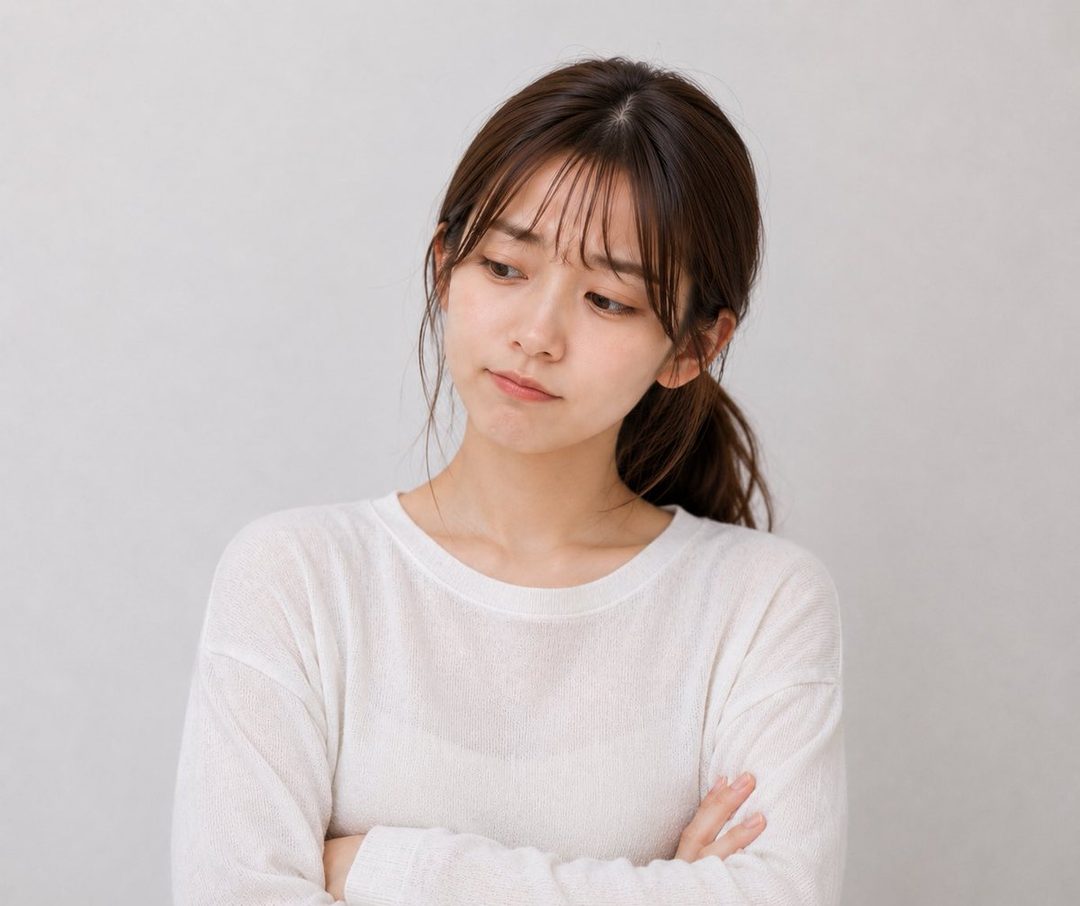 A young East Asian woman with brown hair in a ponytail and bangs, wearing a white long-sleeved top, looks down with a thoughtful expression in a studio setting.