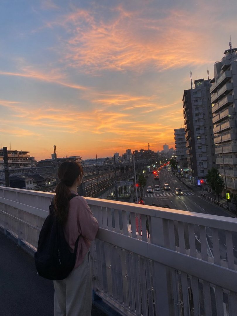 A person with a ponytail and a backpack stands on a bridge, looking out at a city street during a beautiful sunset.