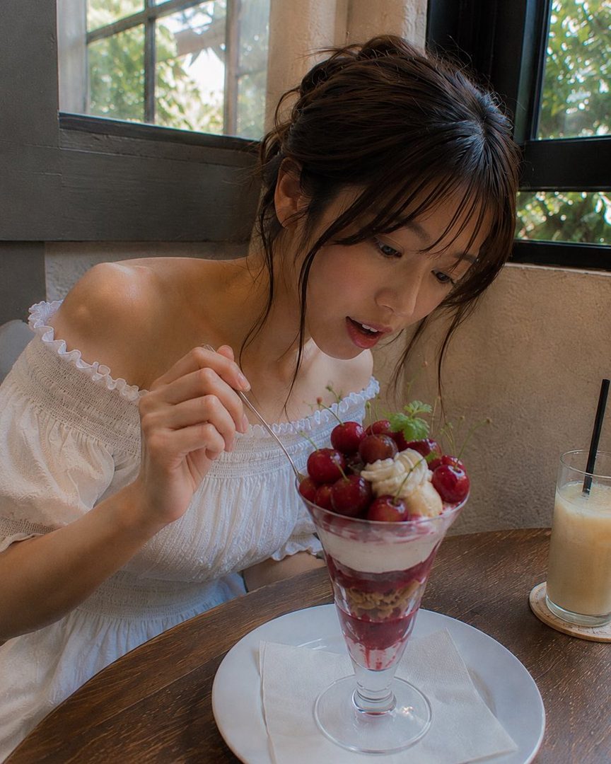 A young woman with brown hair and bangs, wearing a white off-shoulder dress, is enjoying a cherry parfait at a cafe.