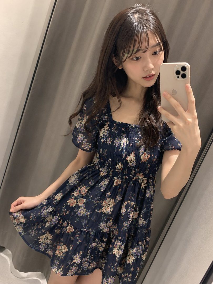 A young woman takes a selfie in a floral dress, looking at her phone.
