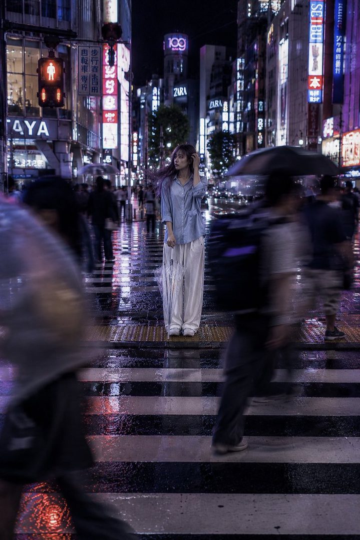 A woman stands on a wet crosswalk at night in a bustling city, surrounded by neon lights and blurred figures.