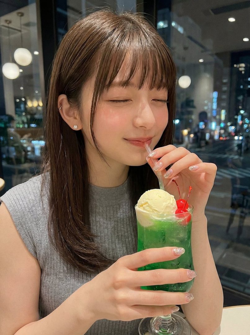 A young woman with brown hair and bangs sips a green cream soda with her eyes closed, in an indoor setting at night.