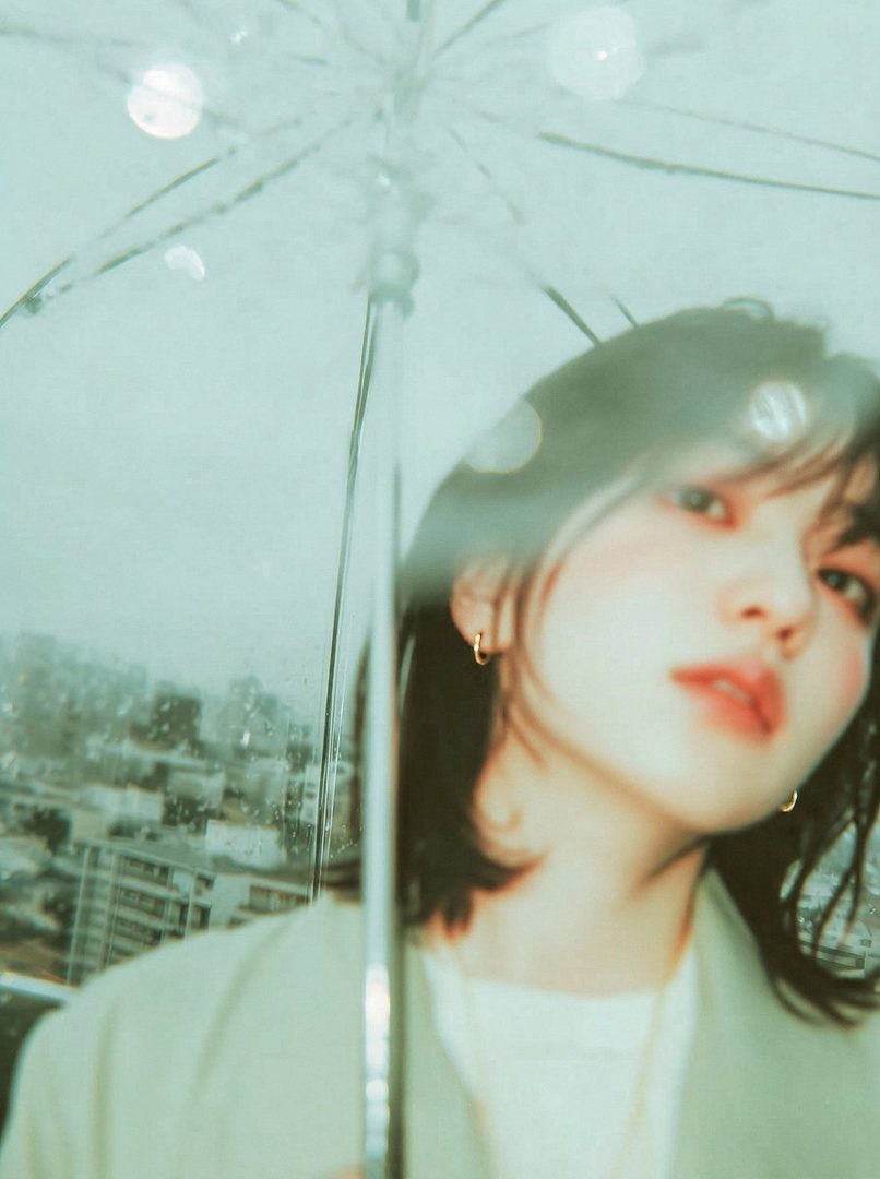 A dreamy portrait of a young woman with short black hair, holding a clear umbrella on a rainy day with a city background.